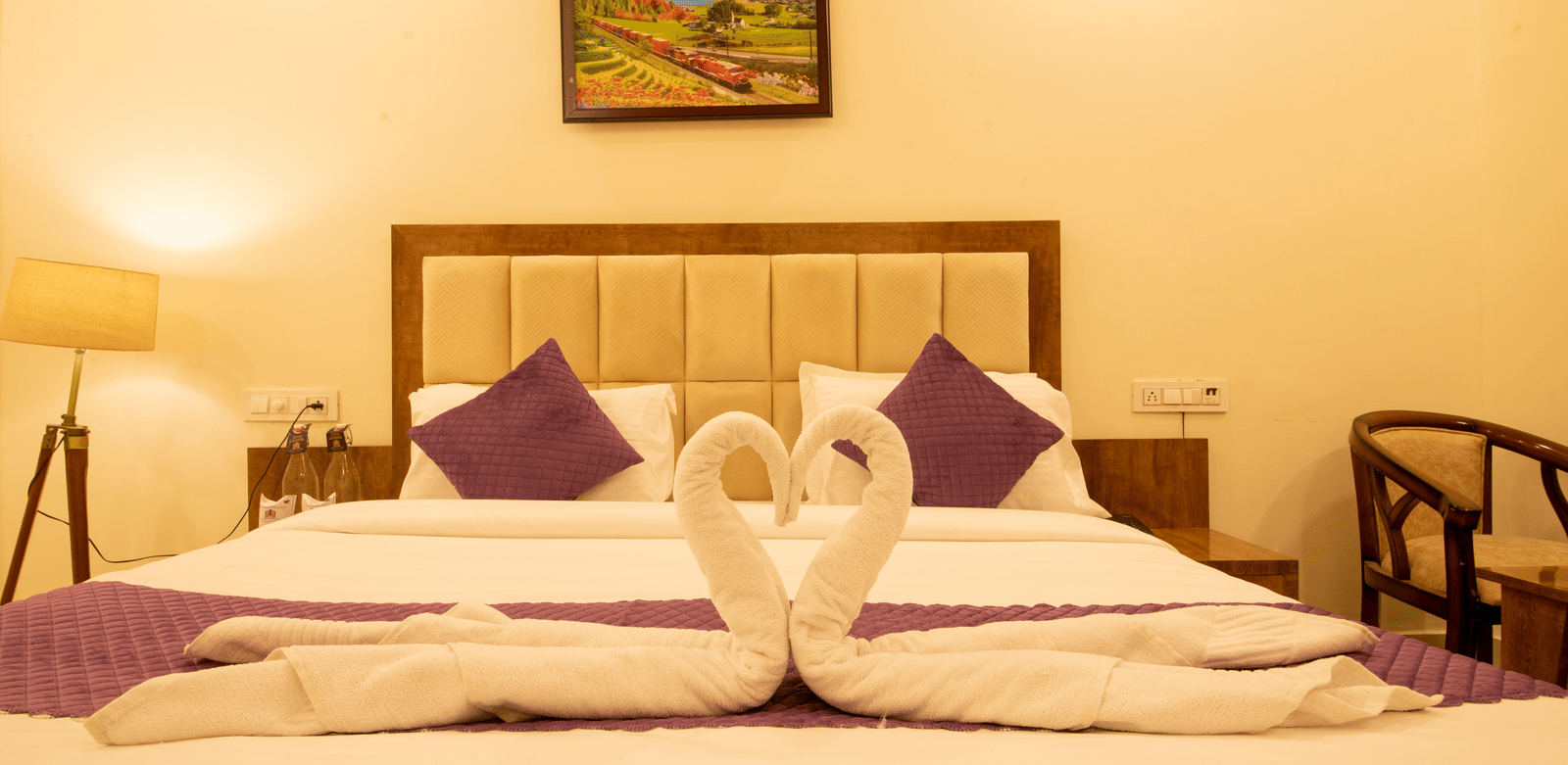 A hotel room, featuring a large bed with a towel swan display, a purple runner and pillows, a wooden headboard, and a painting above the bed | Perfectstayz Value Hills, Rishikesh