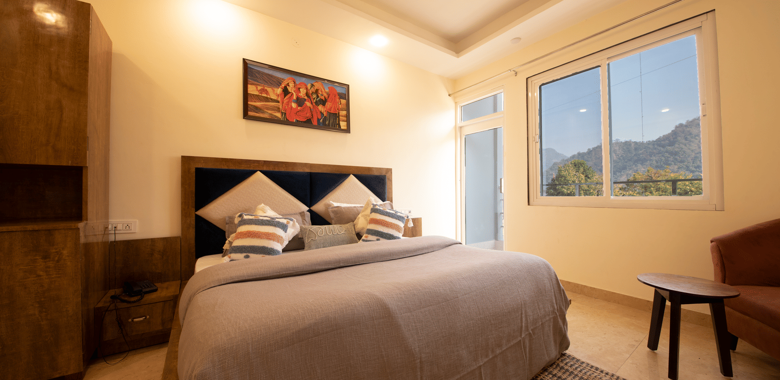 A hotel room, featuring a large bed with a grey duvet and a wooden frame, a painting on the wall, and a window with a view of hills | Perfectstayz Value Hills