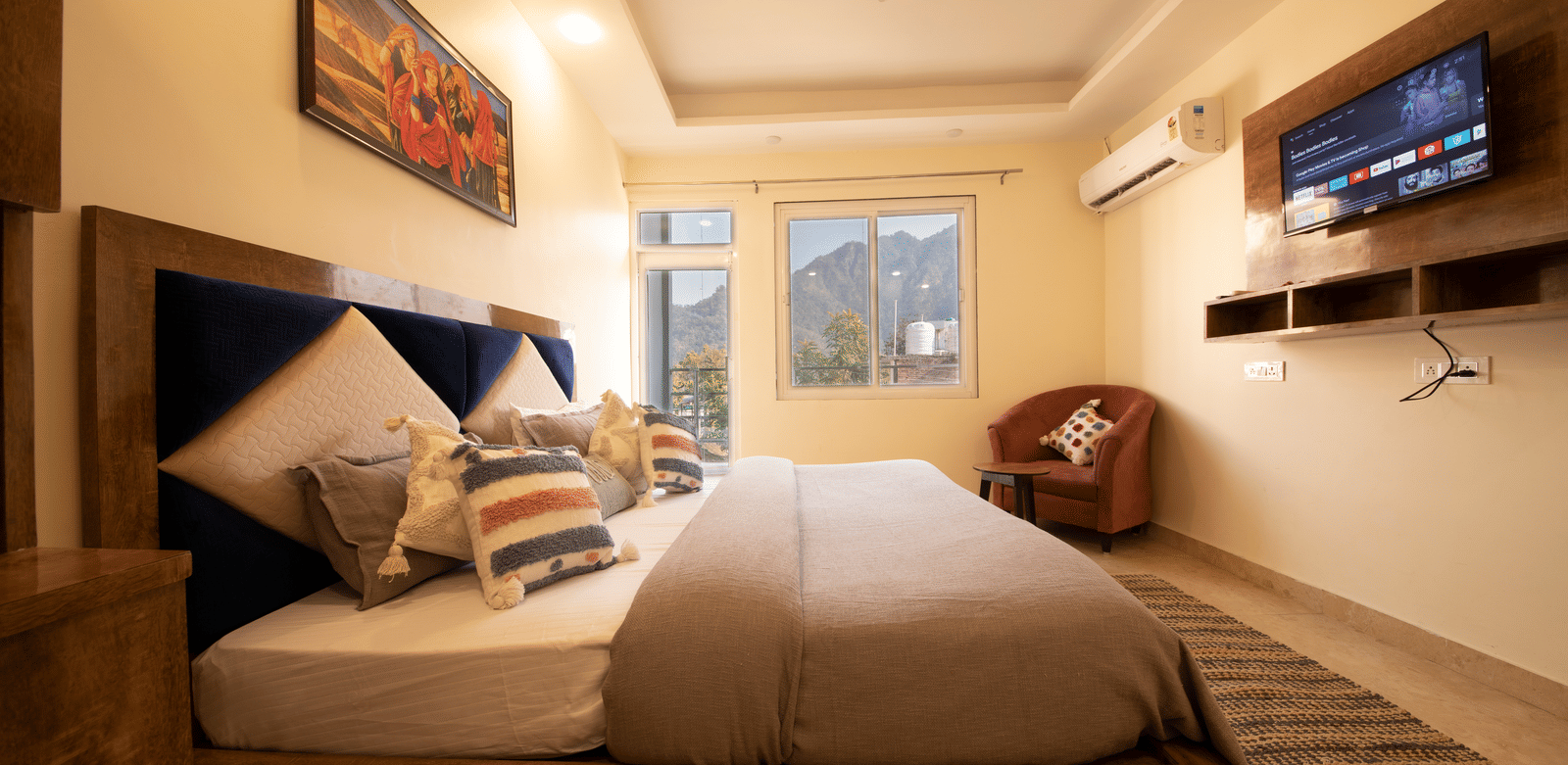 A hotel room, featuring a large grey bed with white linens, blue and orange pillows, a wooden frame, a wall-mounted TV, and a large window with white curtains | Perfectstayz Value Hills, Rishikesh