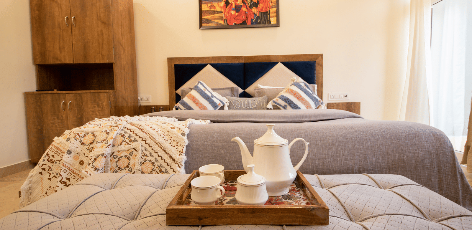 A hotel room, featuring a large grey bed with white linens, blue and orange pillows, a wooden tray with a teapot and cups on the bed, and a painting on the wall | Perfectstayz Value Hills, Rishikesh