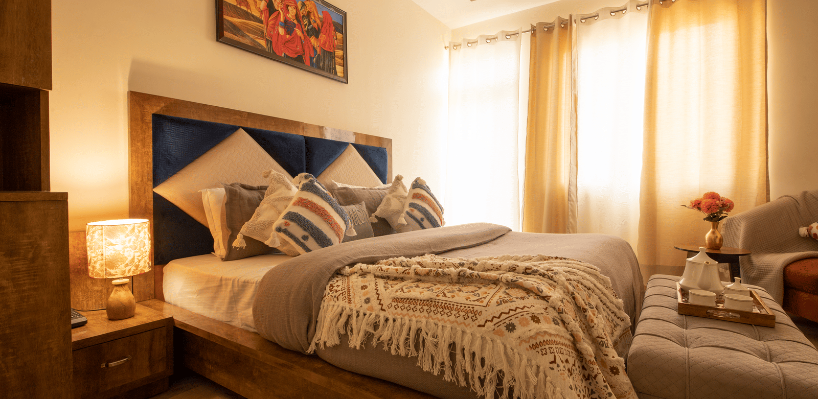A brightly lit hotel room, featuring a large bed with a wooden frame, multiple decorative pillows, a knit throw blanket, and a window with sheer curtains | Perfectstayz Value Hills