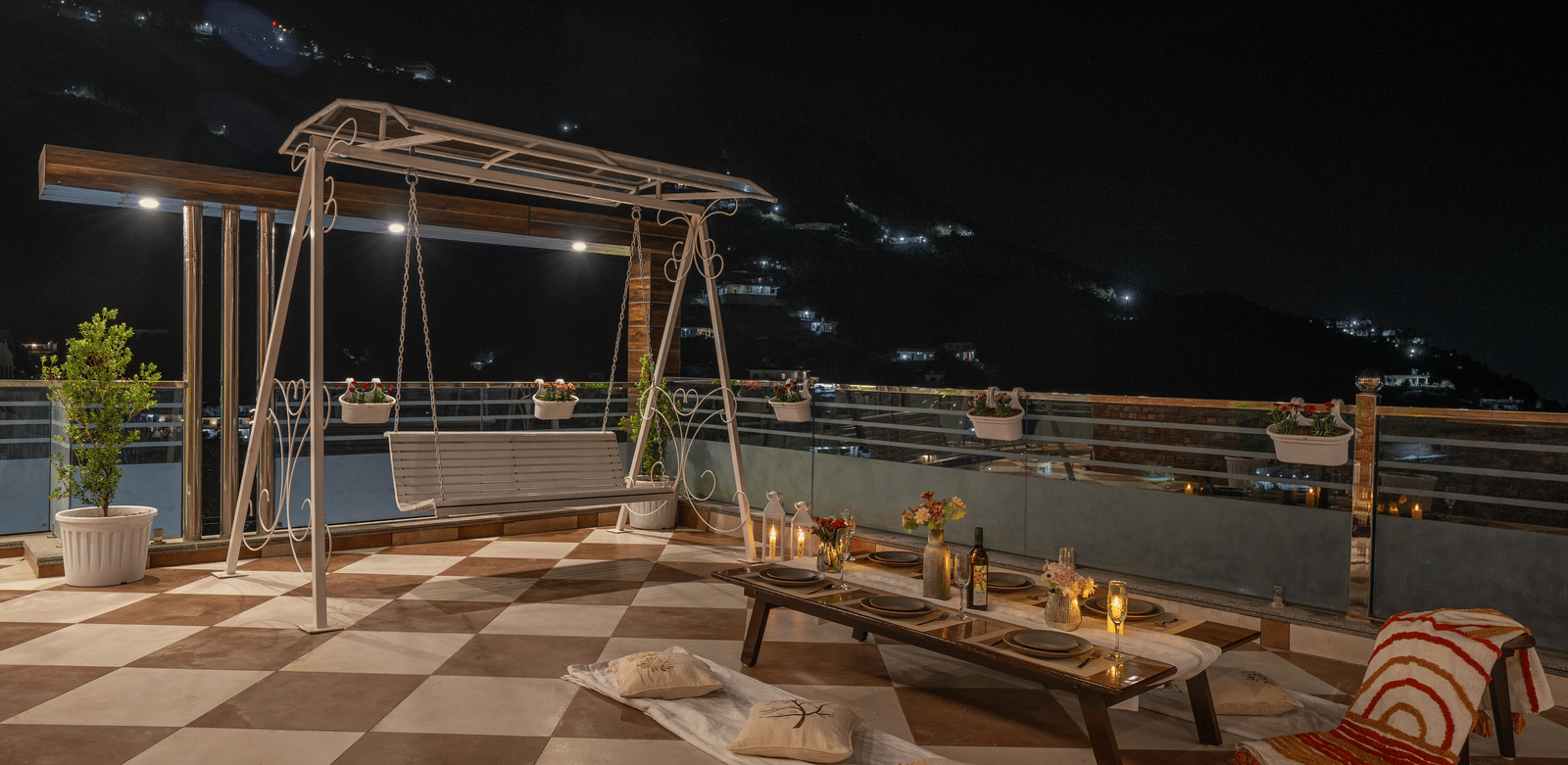 An outdoor terrace at Perfectstayz Value Paradise, Mussoorie with a swing, low table setup with cushions and candles, potted plants and hillside lights in the background at night.