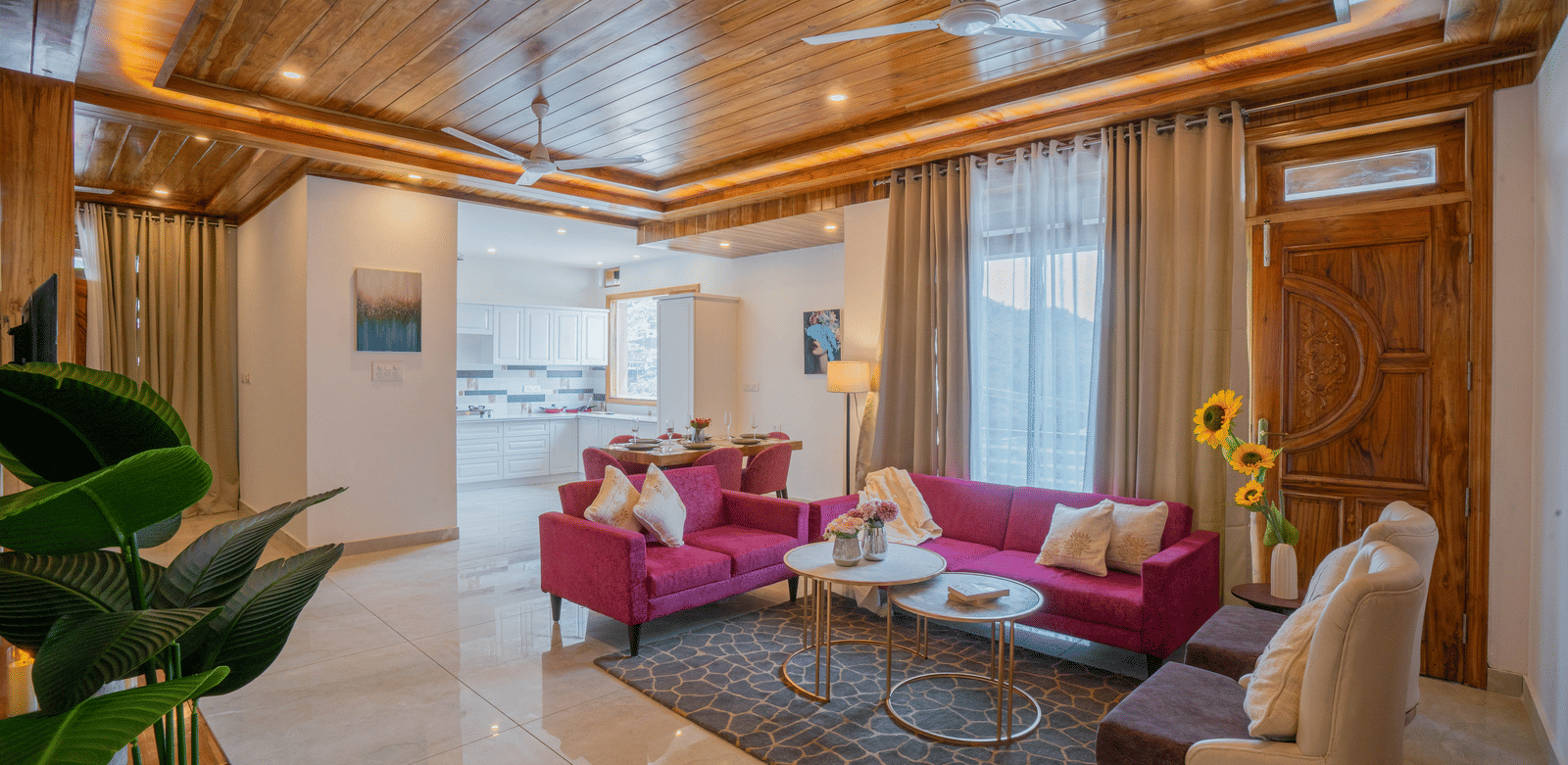 A living room with 2 sofas, a chair, 2 tables, a plant, a wooden ceiling, a dining area, and large windows with curtains at Perfectstayz Value Paradise, Mussoorie.