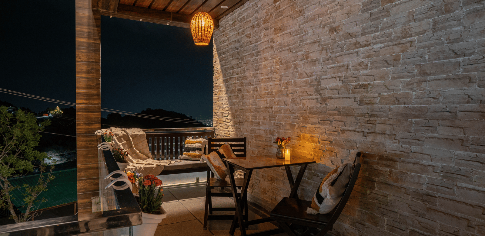 An outdoor seating area with 2 chairs, pillows, a bench, pendant lighting, and a table beside a stone wall at Perfectstayz Value Paradise, Mussoorie.