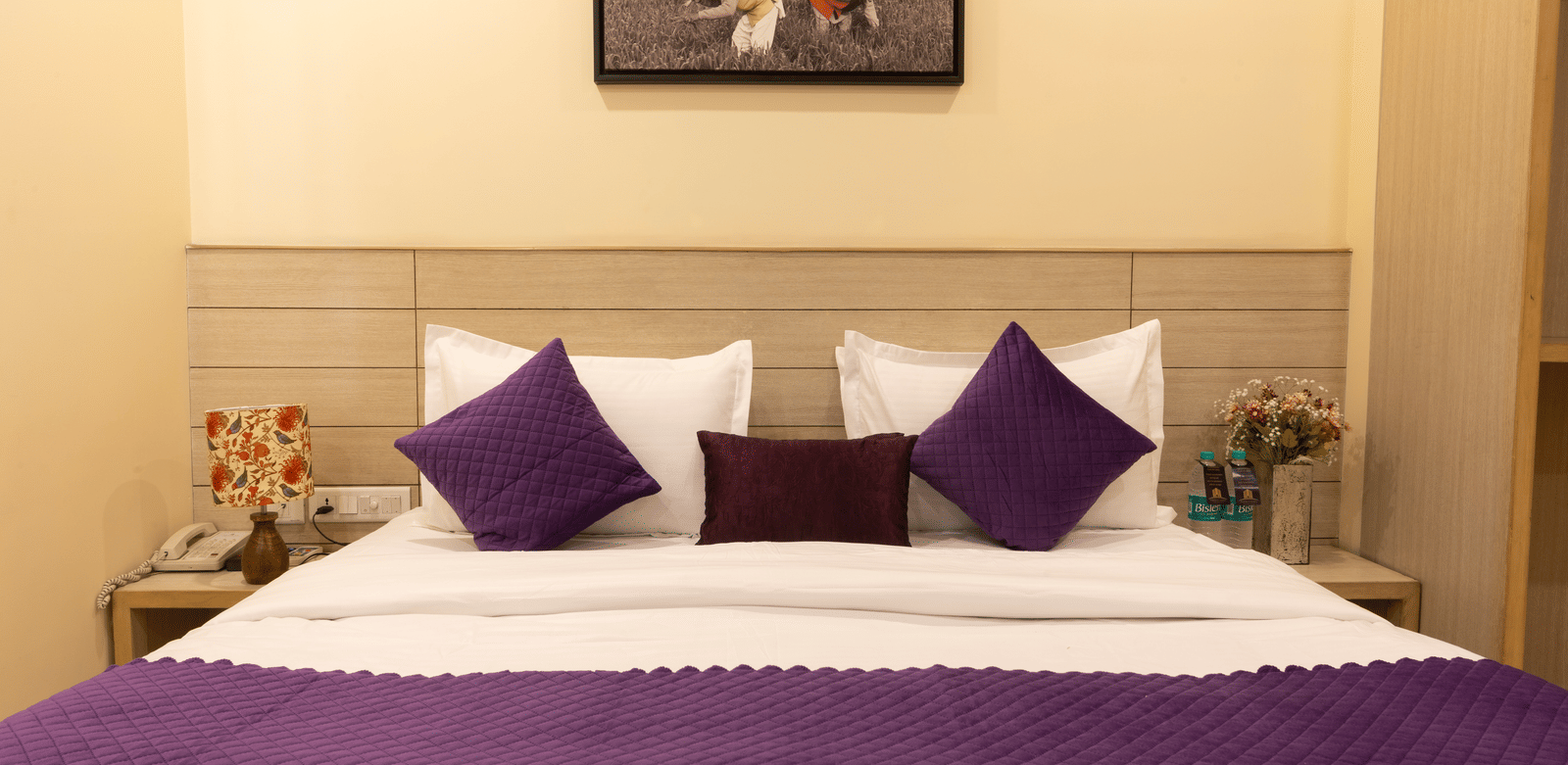 A close-up of a large bed, featuring a white duvet, purple runner and pillows, a light wooden headboard, and a small framed painting on the wall | Perfectstayz Value Sagar