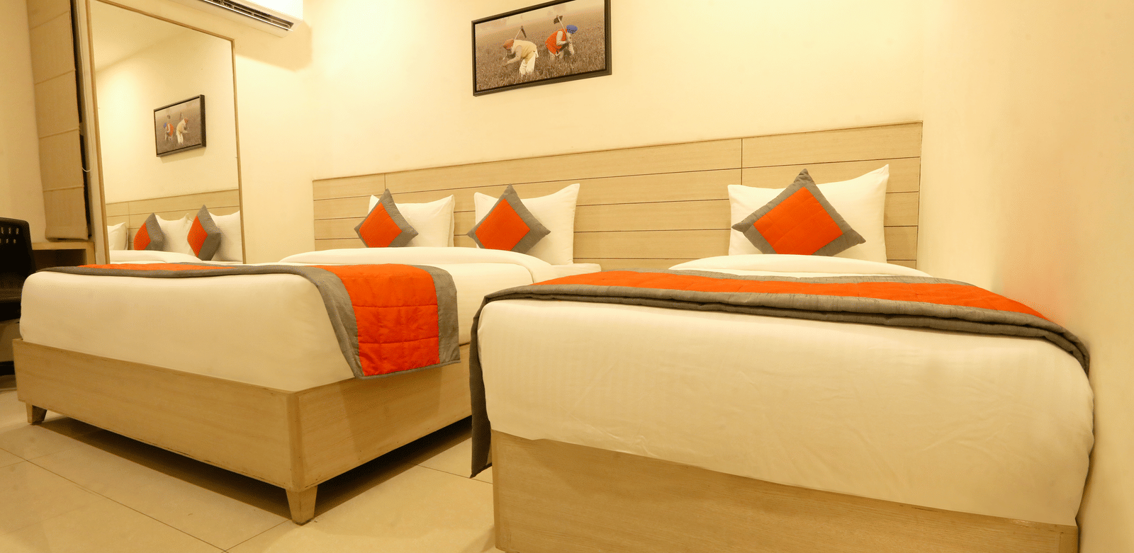 A hotel room, featuring 2 single beds side-by-side with white linens and orange accent runners and pillows, and a painting above the beds | Perfectstayz Value Sagar