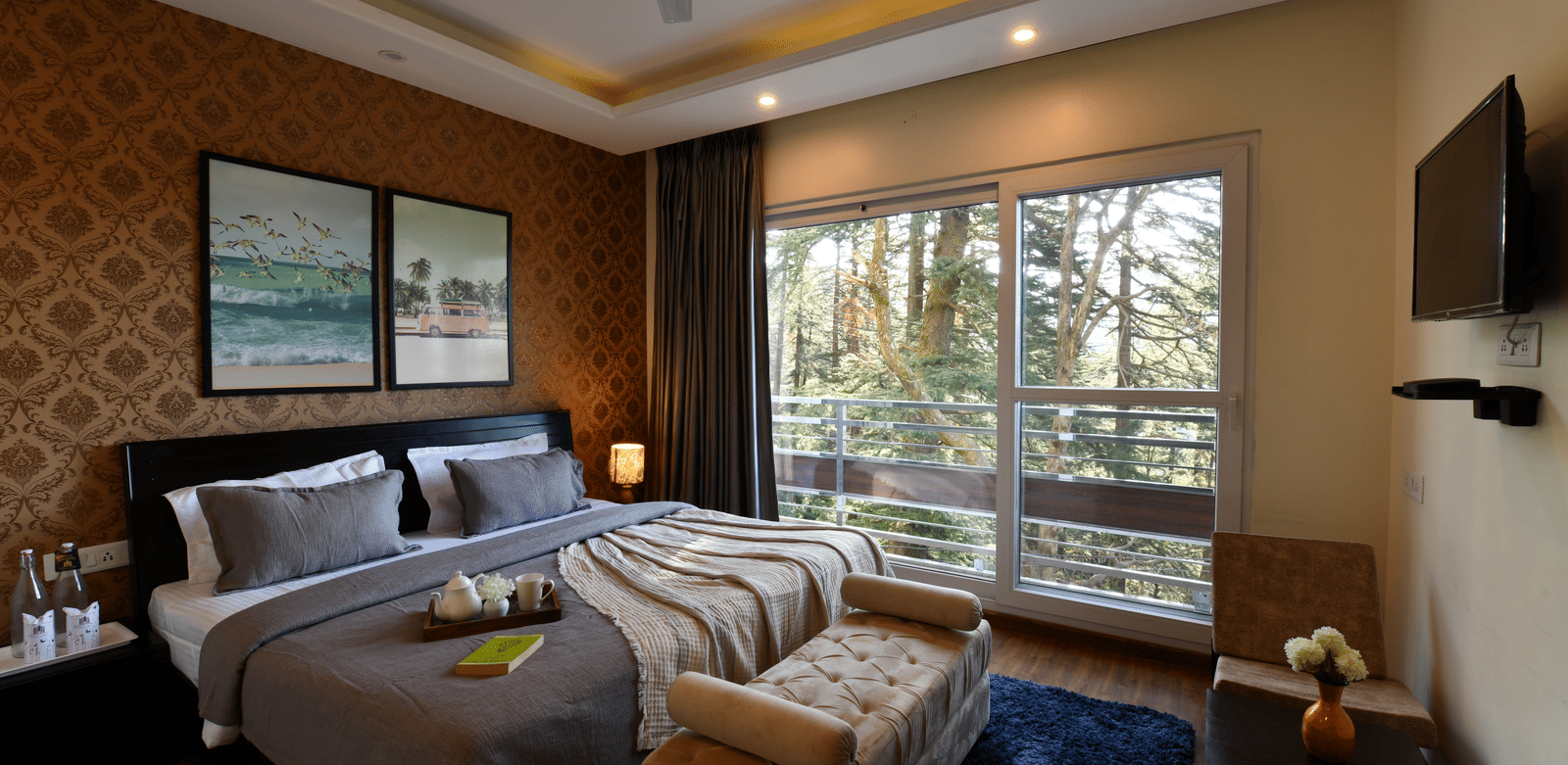 A bedroom at Perfectstayz Value Shimla (Namah Retreat) with a king size bed, a daybed, wall paintings, a television, a tray with cups and kettles, a table, 2 chairs, and a sliding door leading to a balcony.