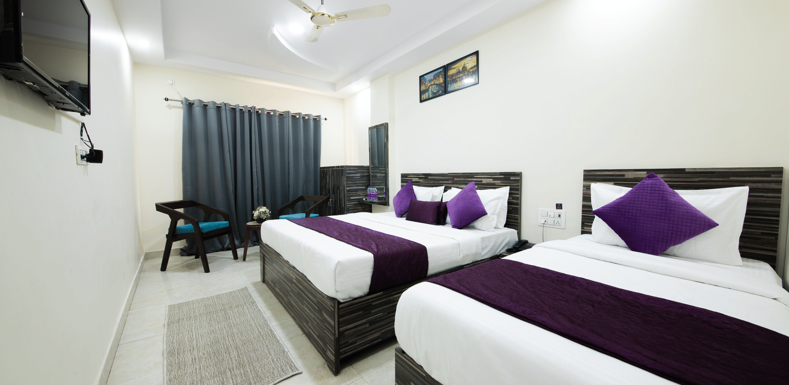 Double bed room with two beds, purple runners, a ceiling fan, seating area, and a TV - Perfectstayz Value Umra Grace, Haridwar