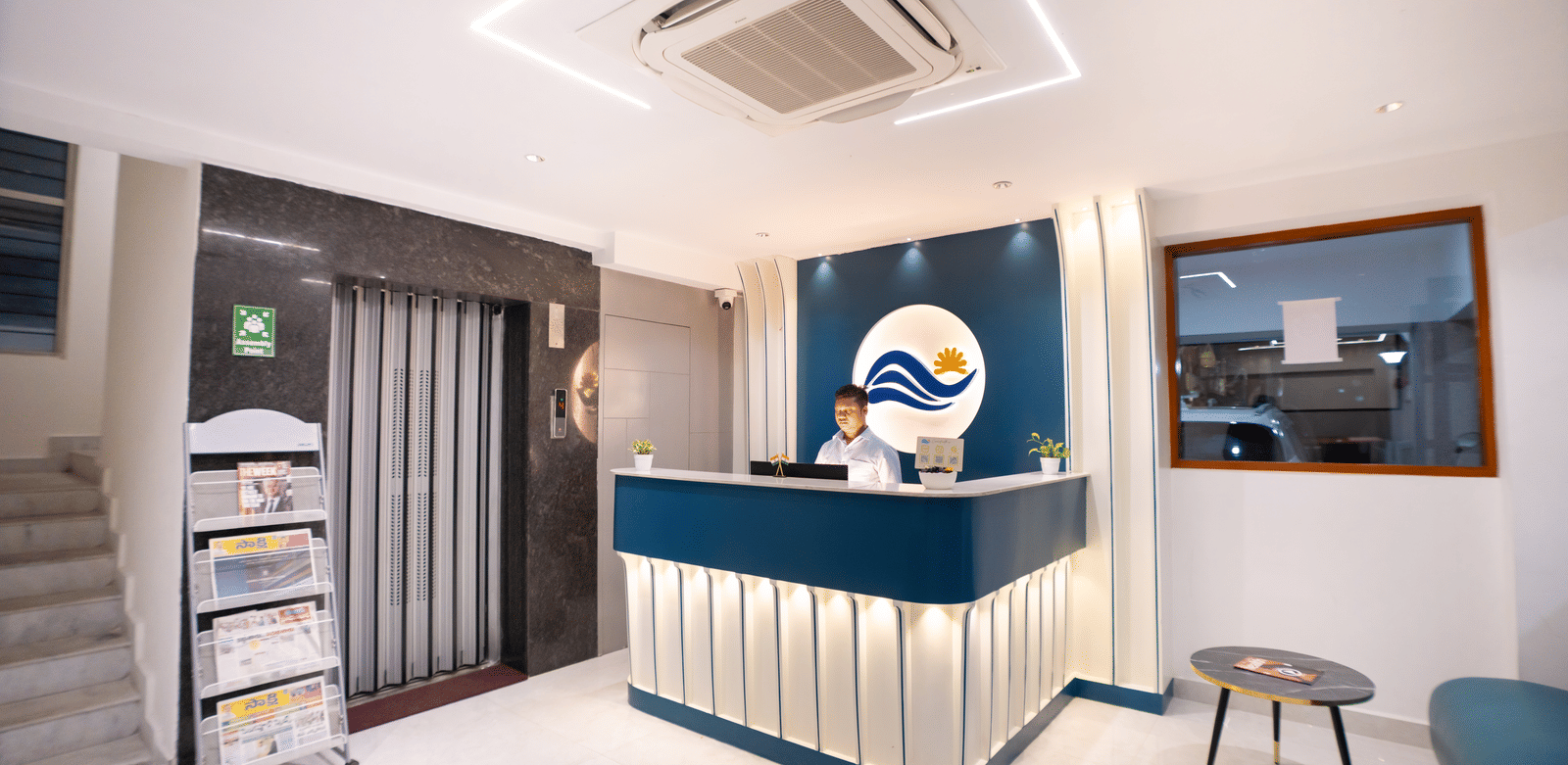 A diagonal view of the reception area with a large desk and a staff member standing behind it, a seating area on one side, and a newspaper stand on the other at Sunrise Business Hotel, Hyderabad.