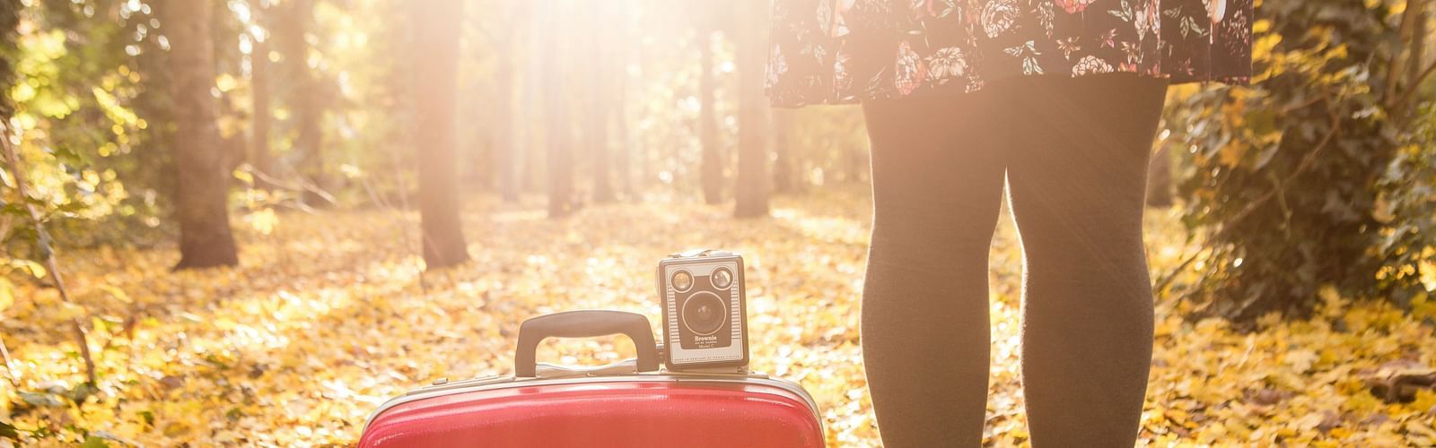 A close up shot of a woman standing next to a red suitcase in a forest