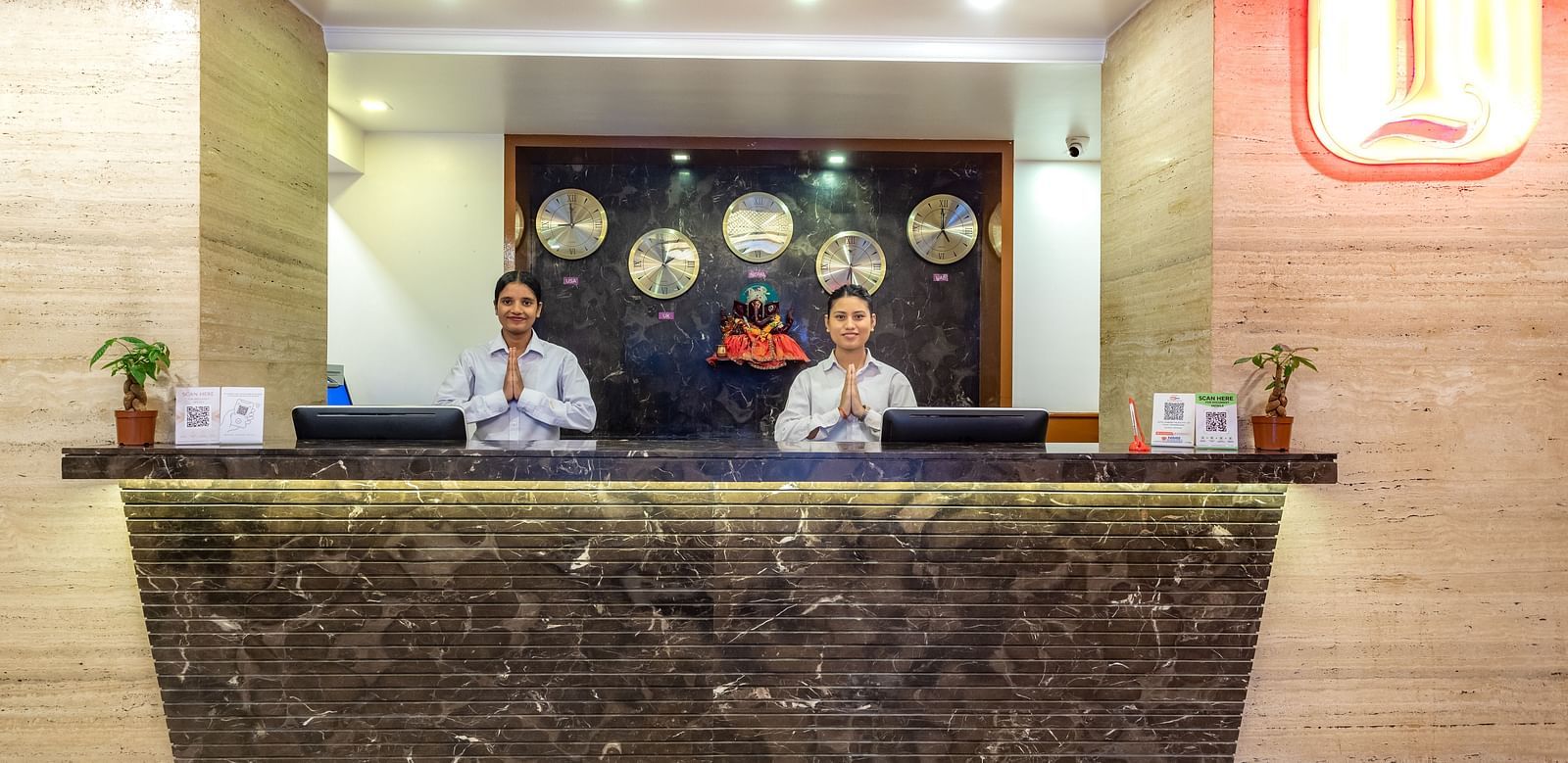 Hotel reception at Diamond Palace Lords Plaza with two staff members behind a marble counter and world clocks on the wall.
