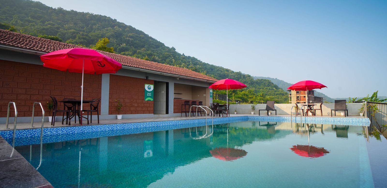 A swimming pool with red umbrellas and lounge chairs, set against a backdrop of mountains and a building - Gaurav Lords Resort Shrivardhan.