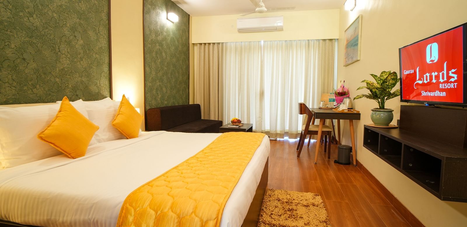 A hotel room with a bed, a television featuring the Lords logo, a dining table, a sofa, a rug, and a plant - Gaurav Lords Resort Shrivardhan.