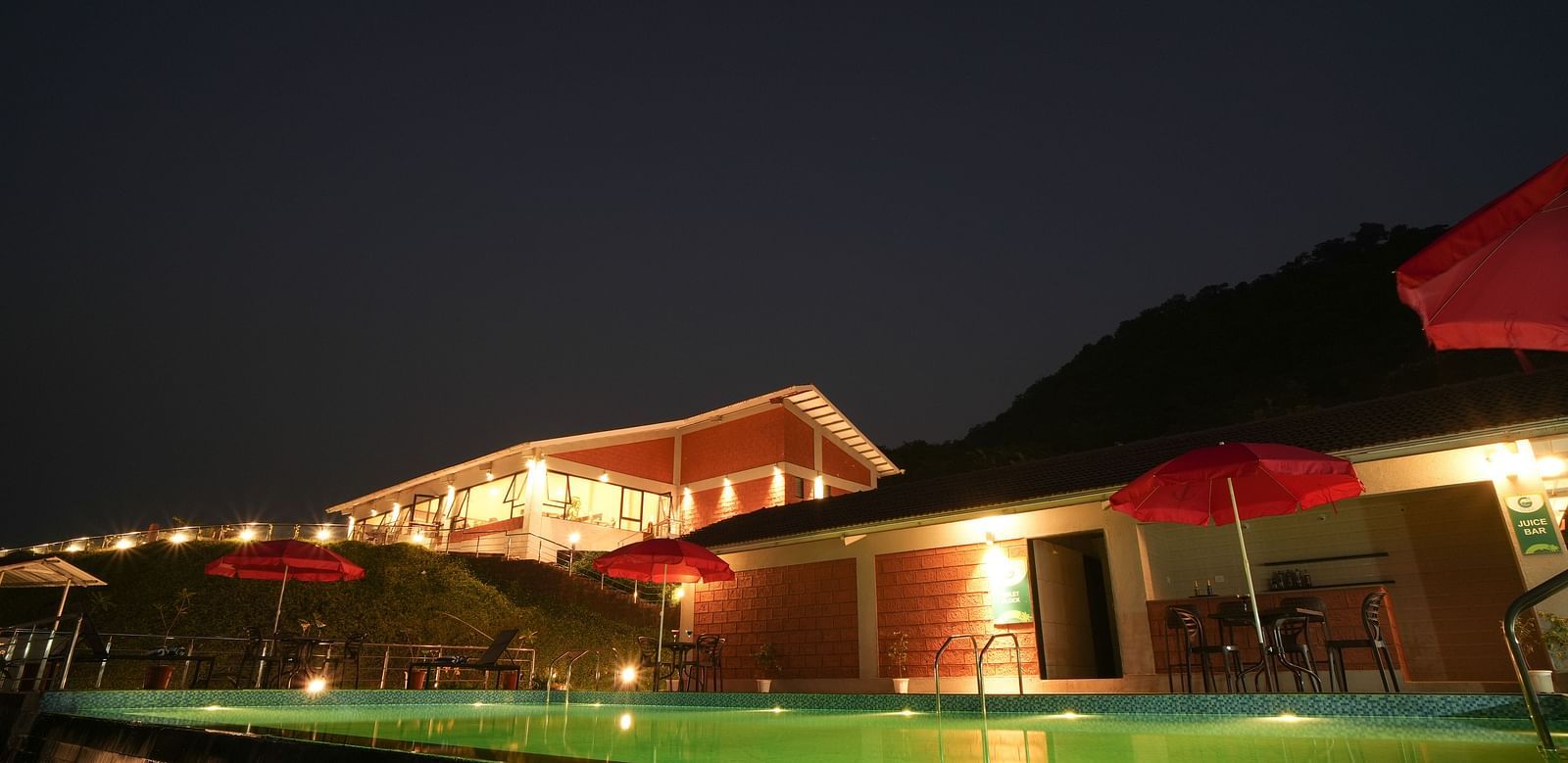 A swimming pool lit at night with red umbrellas and a building in the background - Gaurav Lords Resort Shrivardhan.