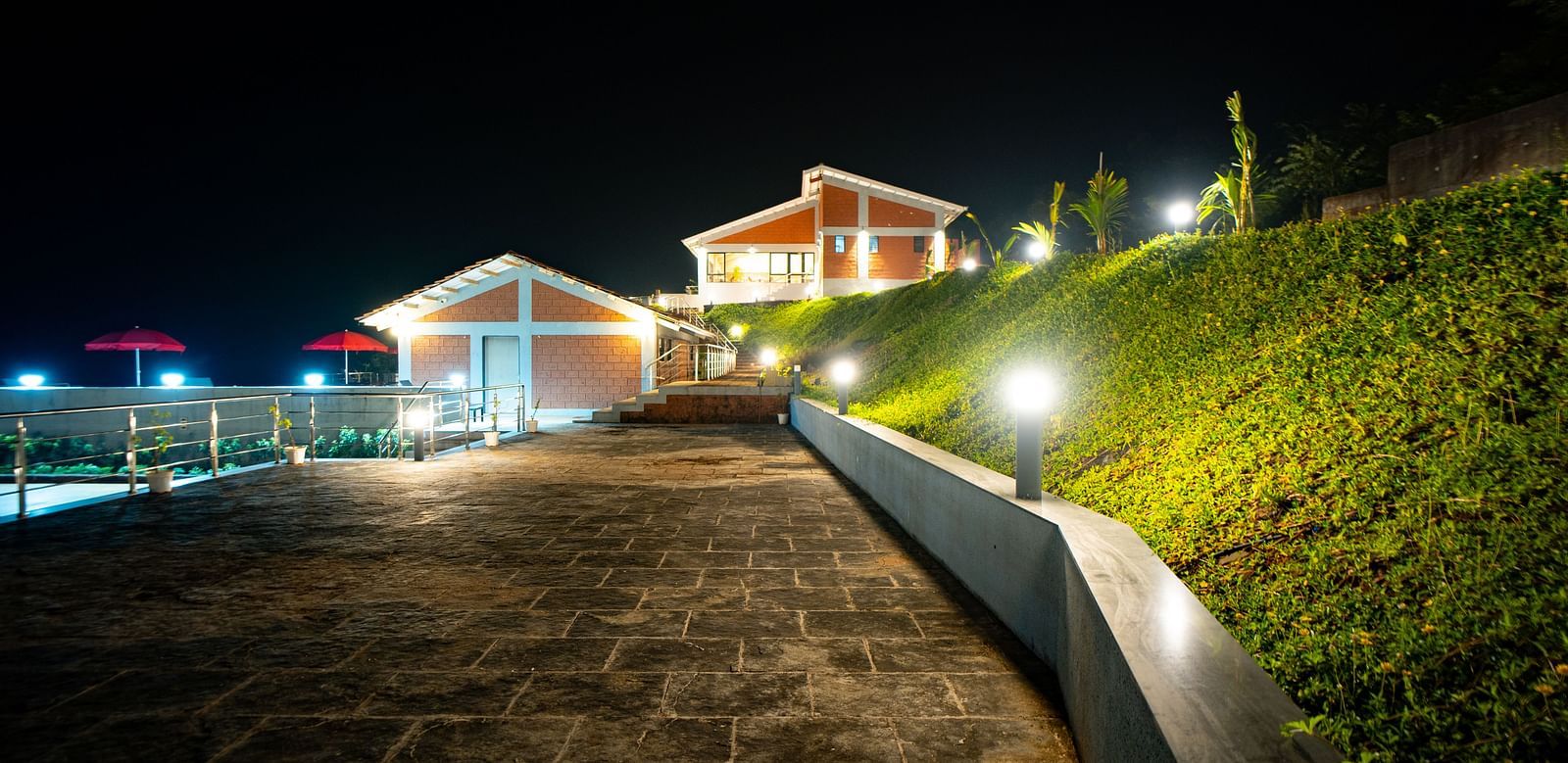 A stone pathway leading to illuminated buildings at night, with a green hillside on the right - Gaurav Lords Resort Shrivardhan.