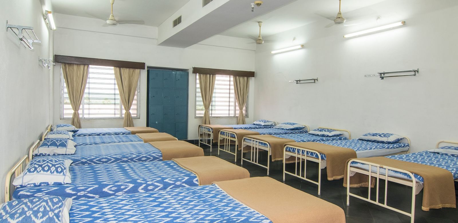 Beds with bedding are arranged in two rows in a room with two windows at Hotel Sahara - Comfortable Dormitory in Ramoji Film City, Hyderabad.