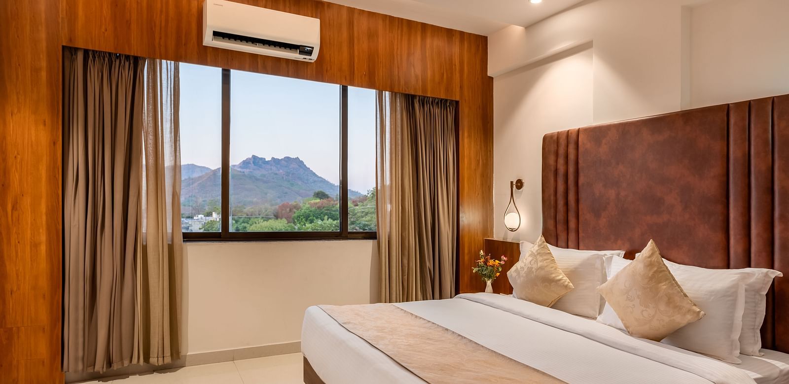 A comfortable king-size bed with soft pillows and a throw faces a window with curtains revealing a mountain view - Lords Eco Inn Ambaji.