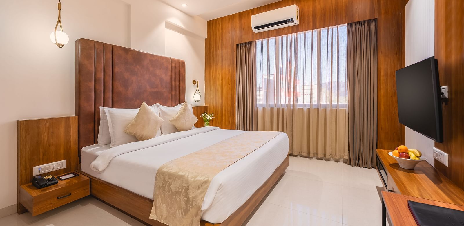 A comfortable king-size bed with soft pillows and a decorative throw faces a mounted television in a well-lit hotel room with a window - Lords Eco Inn Ambaji.