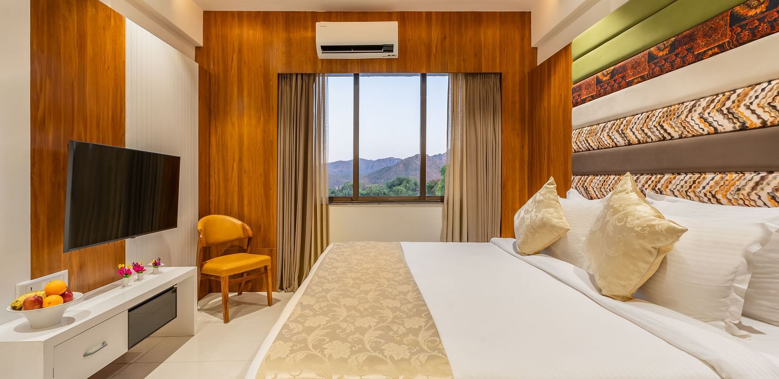 A well-appointed hotel room features a king-size bed, a mounted television, a cushioned chair, and a view through the window - Lords Eco Inn Ambaji.