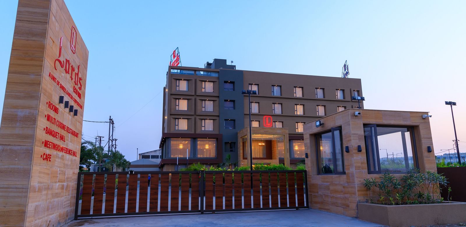The exterior of Lords Eco Inn Apple Gate is viewed from the entrance, showing the multi-storey building, signage, and surrounding gates under a clear sky.