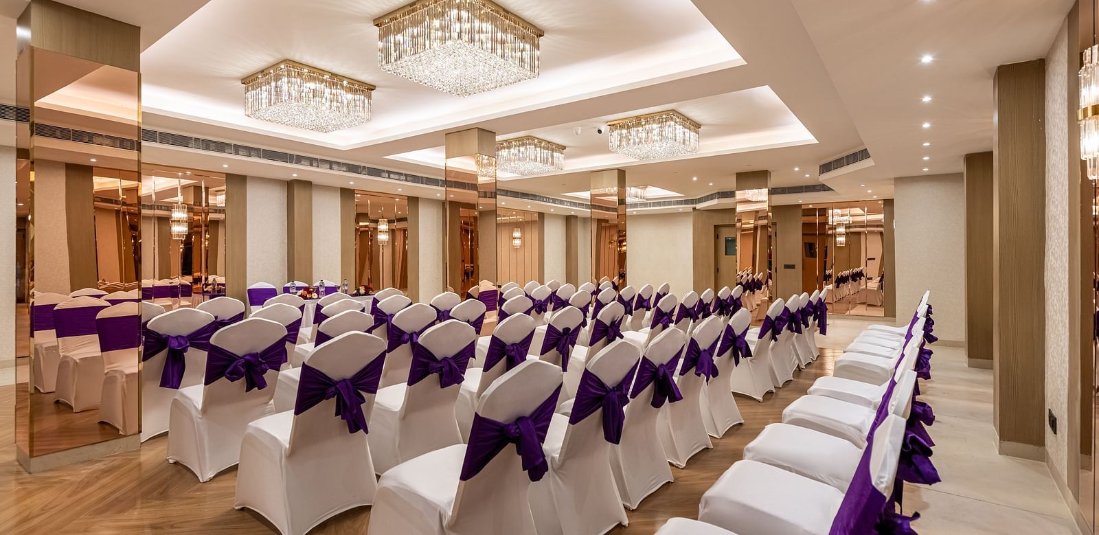 An event room with rows of white chairs adorned with purple sashes. It features a decorative ceiling with several chandeliers and mirrored wall panels - Lords Eco Inn Dehradun.