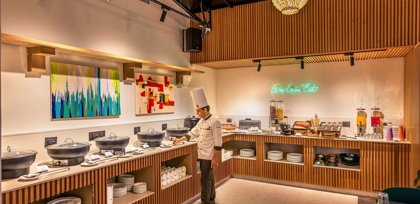 A buffet area with a chef standing near serving dishes, offering a variety of food and drinks. It features wooden paneling, a chandelier, and colourful artwork on the wall - Lords Eco Inn Dehradun.