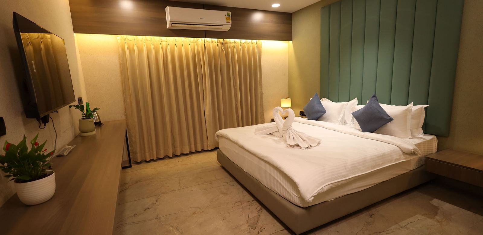 The image shows a well-lit bedroom with a bed, pillows, bedside lamps, curtains, and a dresser with plants - Lords Eco Inn Navsari.