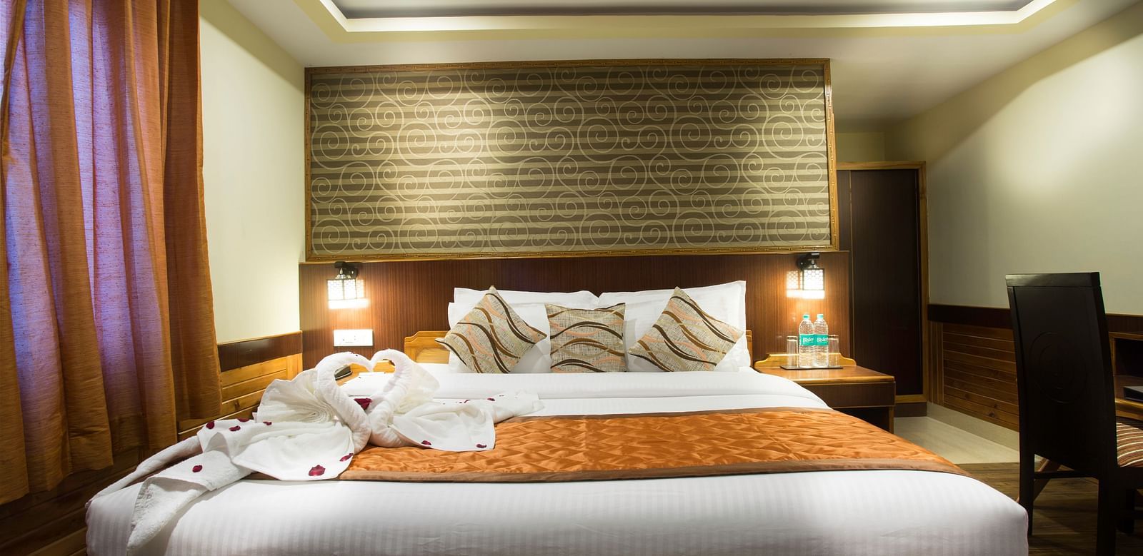A room at New Orchid Lords Inn Gangtok contains a bed, pillows, folded towels, bedside tables, lamps, curtains, and a desk with a chair.