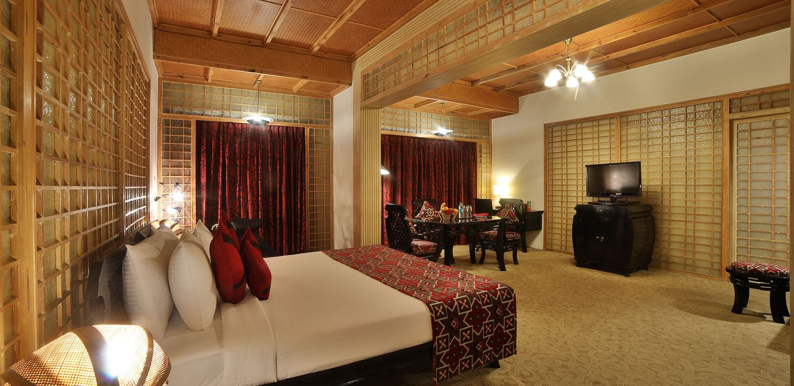 A warmly lit hotel room with traditional decor, a large bed, and wooden lattice work adding to the ambiance A grand hotel suite with an opulent red and white decor, large bed, and seating area, reflecting a traditional style at Sitara Luxury Hotel in Hyder