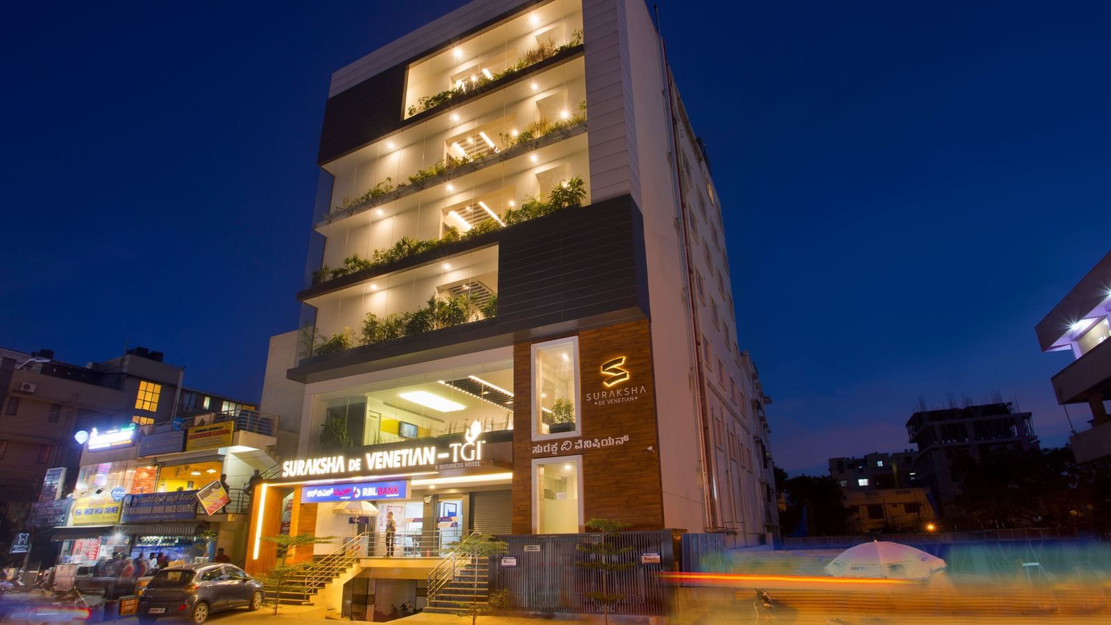 An exterior view of a modern multi-story building illuminated at dusk with signage and street activity - De Venetian by TGI, Brookefield