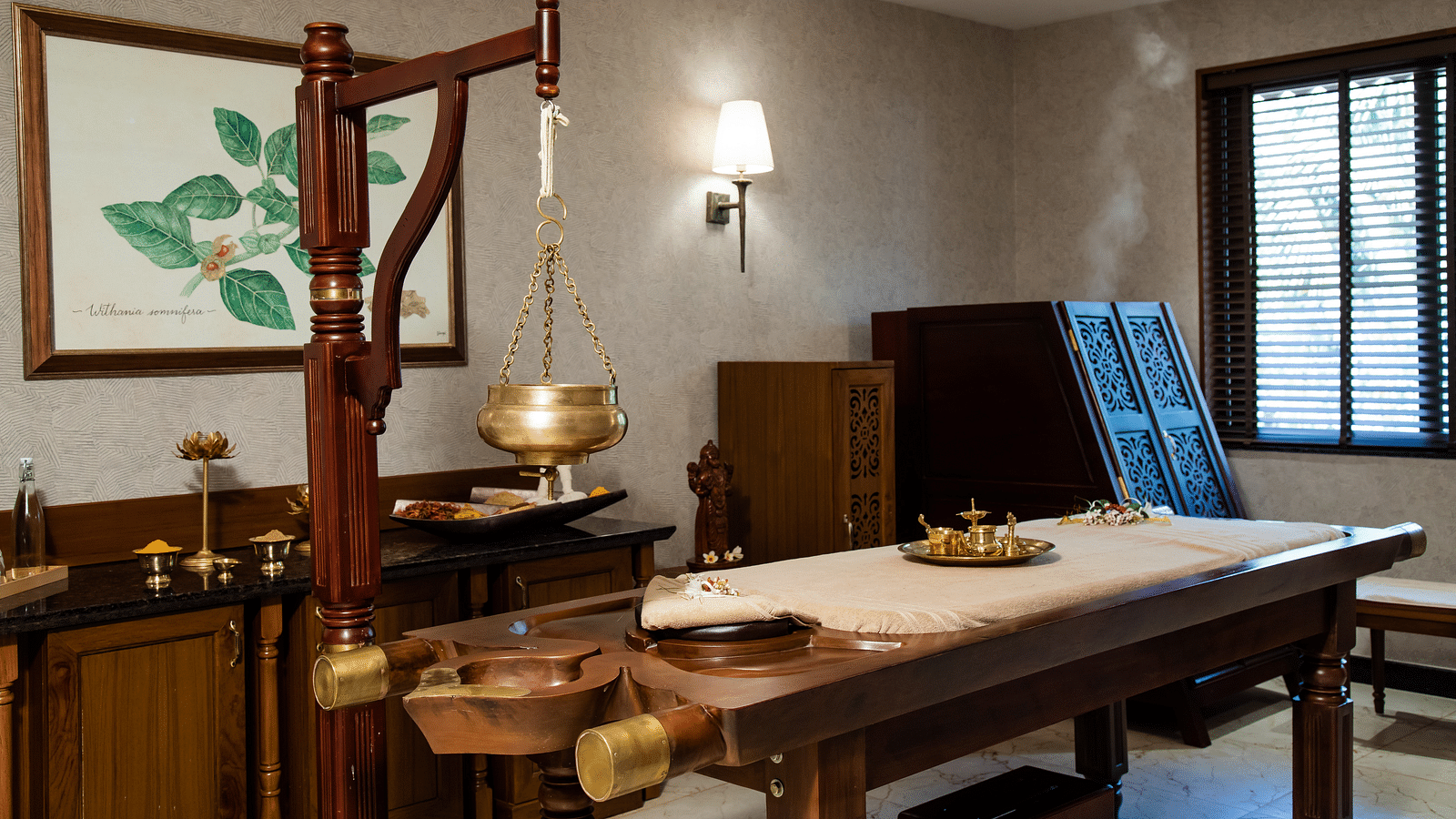 An Ayurvedic treatment room with a wooden massage table and hanging brass pot - Gamyam Retreat