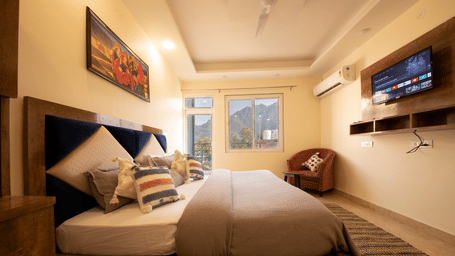 A hotel room, featuring a large grey bed with white linens, blue and orange pillows, a wooden frame, a wall-mounted TV, and a large window with white curtains | Perfectstayz Value Hills, Rishikesh
