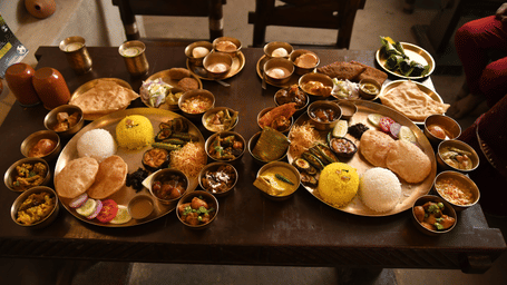 A traditional Bengali thali spread at Vedic Village Spa Resort featuring multiple brass bowls and plates filled with various dishes arranged on a long wooden dining table.