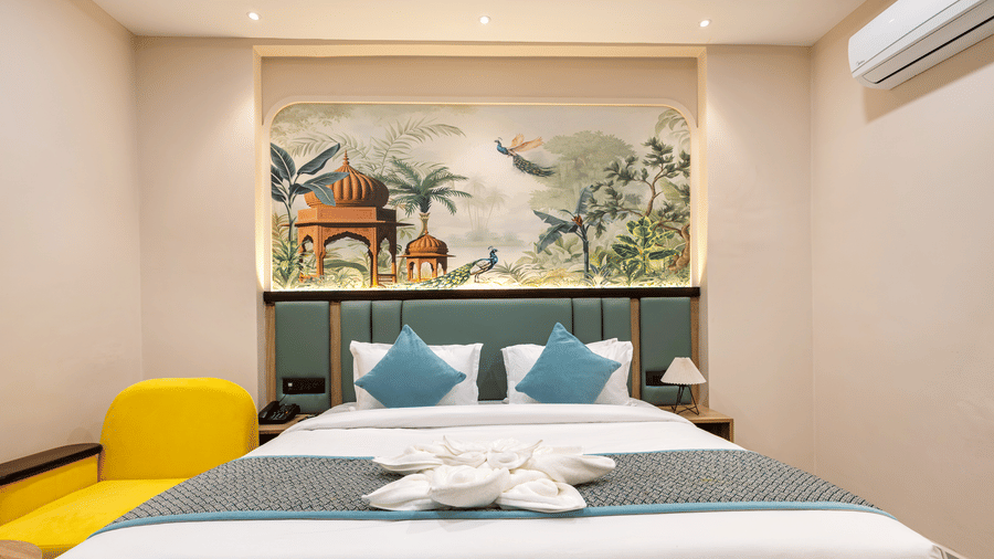 A bedroom with a king size bed made with white linens and blue pillows alongside a yellow chair and an air-conditioner, at Monday Hotels & Suites Madhapur Hyderabad.