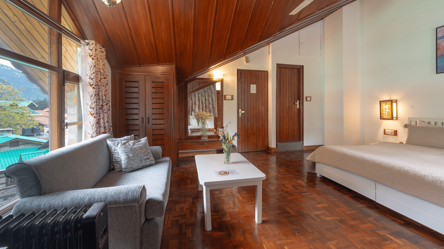 A premium room with a slanted wooden ceiling, featuring a grey sofa, coffee table, and large window view - Johnson Lodge & Spa, Manali