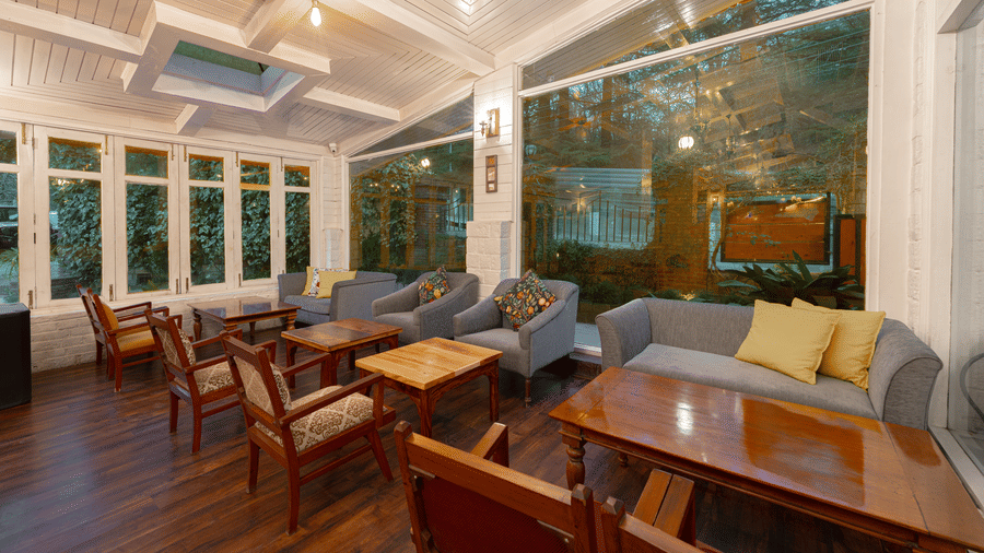 A sunlit veranda cafe with large glass windows overlooking the property’s trees, featuring comfortable grey sofas - Johnson Lodge & Spa, Manali