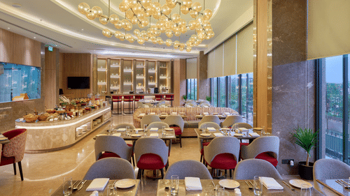 A restaurant interior at KPG Sarovar Portico, Raj Nagar Extension, Ghaziabad featuring dining tables, chairs, a decorative ceiling fixture, and large windows.