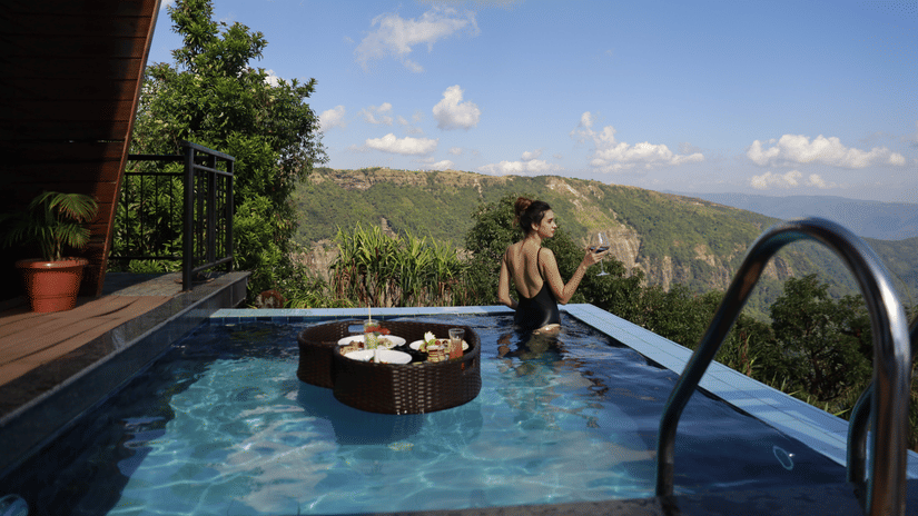 Woman in black swimsuit with floating breakfast tray and red wine in private plunge pool at Polo Resort Cherrapunji overlooking the Khasi Hills.