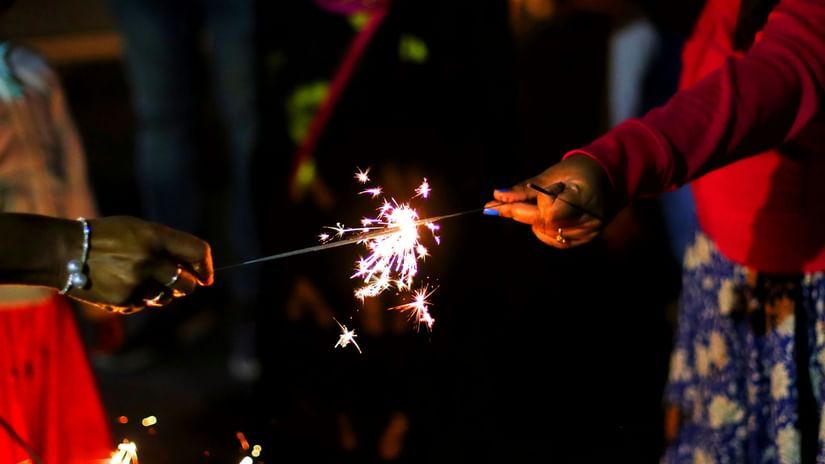 Two people, one on the left and one on the right, are holding and lighting a handheld sparkler. The sparkler emits a spray of bright, glowing sparks against a dark background.