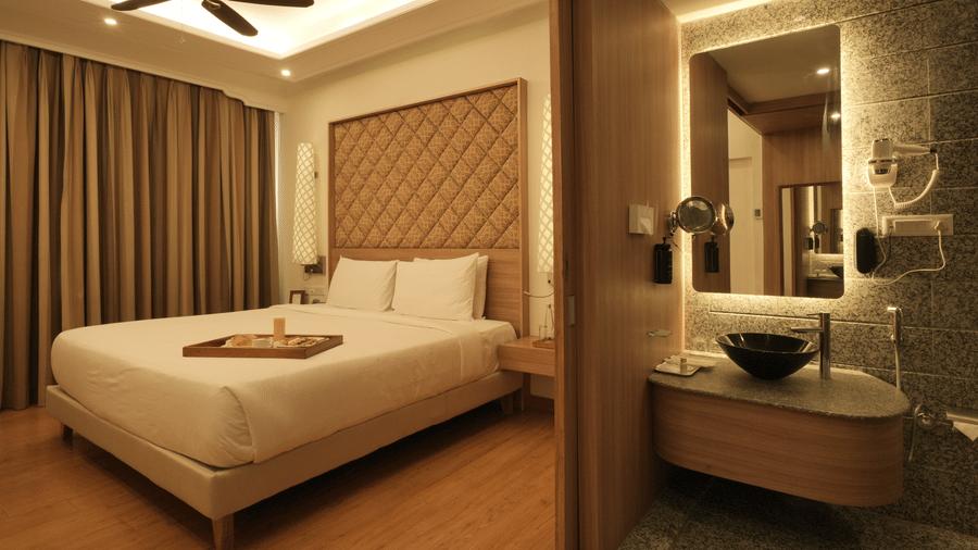 A spacious bedroom with a king size bed, upholstered headboard, wooden flooring, and an attached open bathroom with a glass partition in one of the best hotel in Jaipur - Clarks Amer, Jaipur