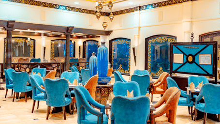 The main dining area of Za-aiqa, located within Noor-Us-Sabah Palace, Bhopal, featuring vibrant teal velvet chairs, arched wall alcoves, and a spacious, elegant layout.