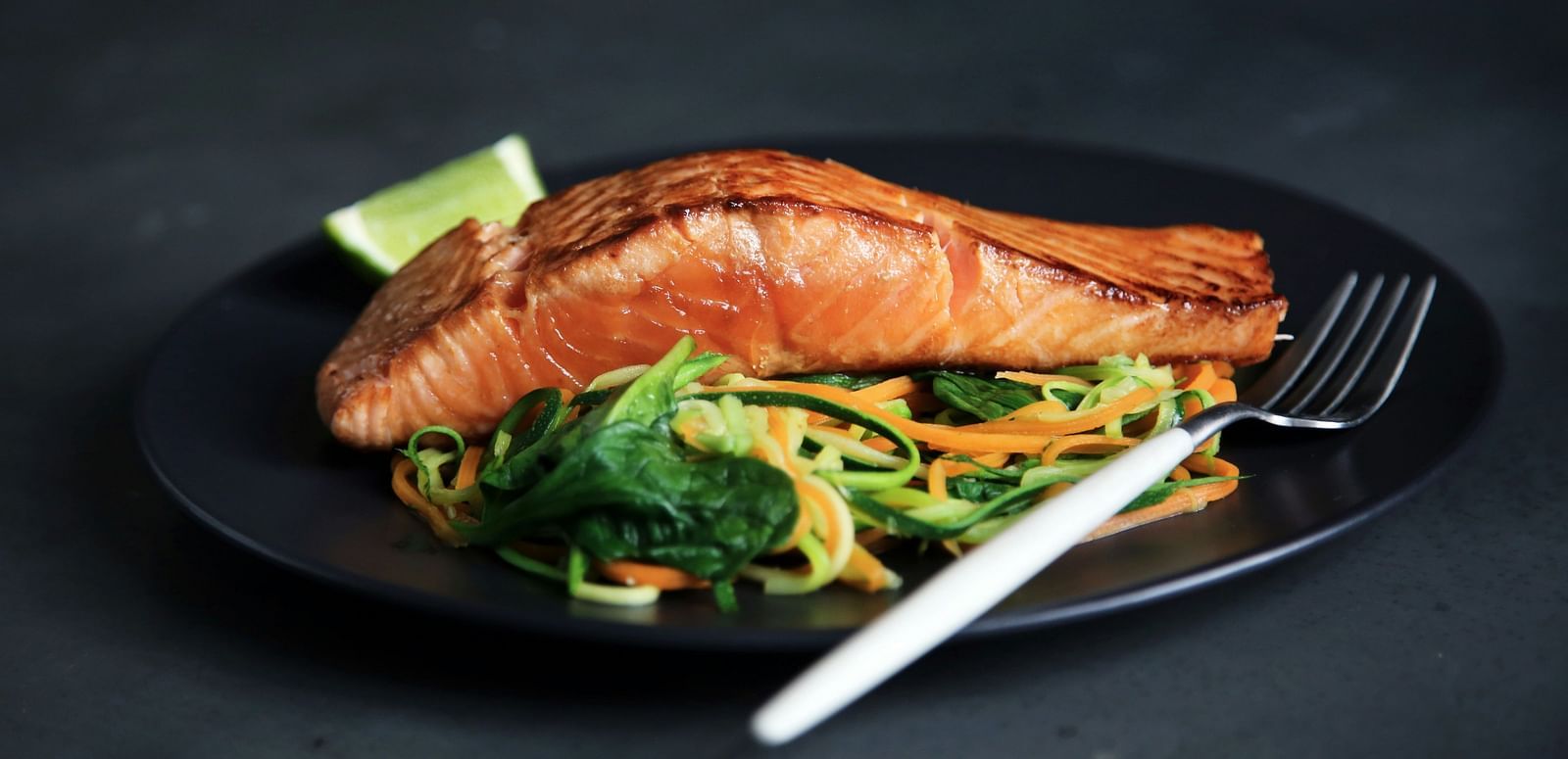 A plated dish of grilled salmon served on a bed of sautéed greens and noodles, with a lime wedge on the side.