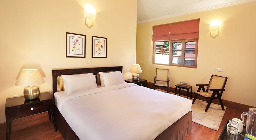 An image of a deluxe room with a double size bed, bedside table with lamps on either side, a chair, and other decorative items - Lamrin Boutique Cottages, Rishikesh