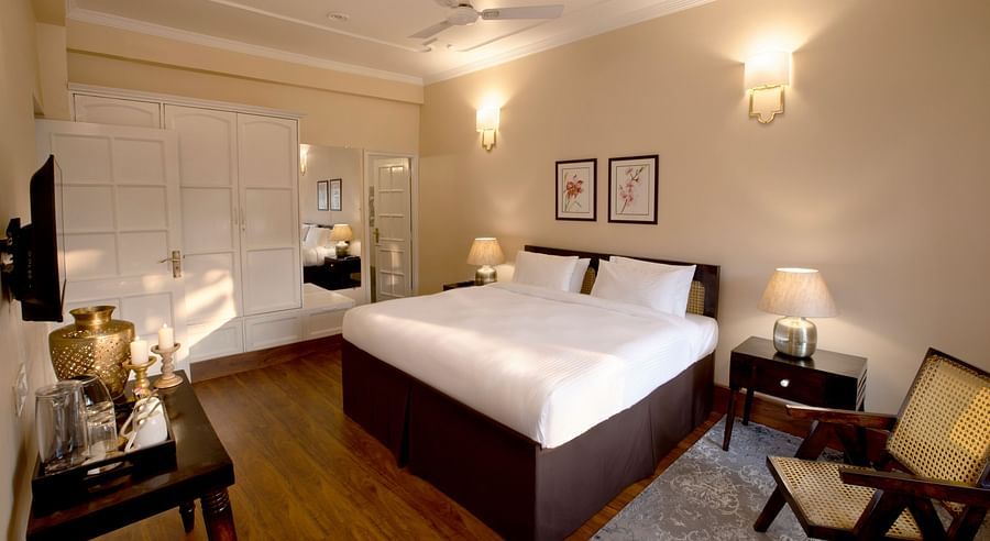 An image of super deluxe room featuring a king size bed, bedside table on either side, a flat TV screen, a table where antique items are placed, and  a chair in one corner of the room- Lamrin Boutique Cottages Rishikesh