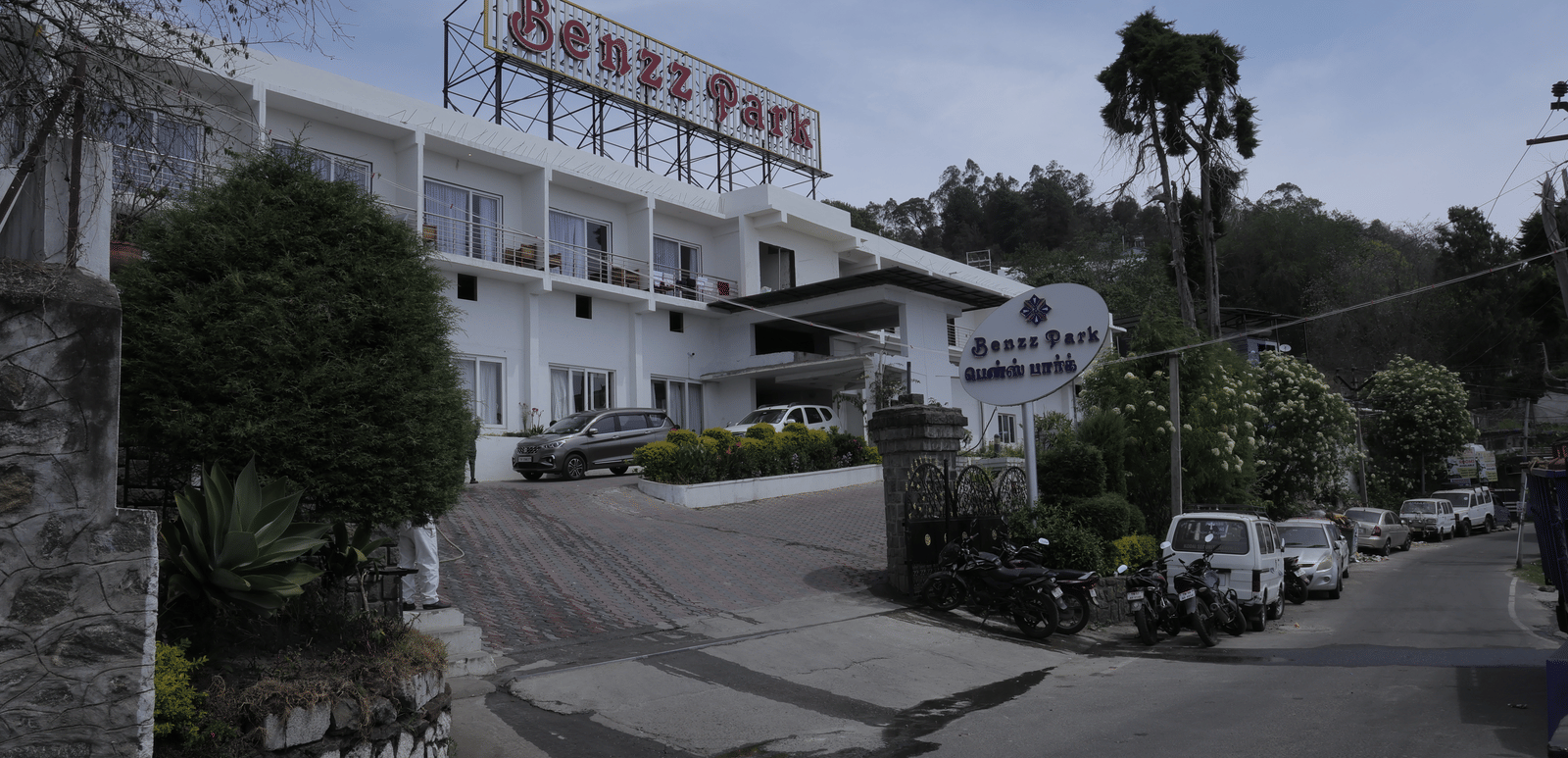 The Benzz Park hotel building exterior on a hillside road, surrounded by trees and vehicles.