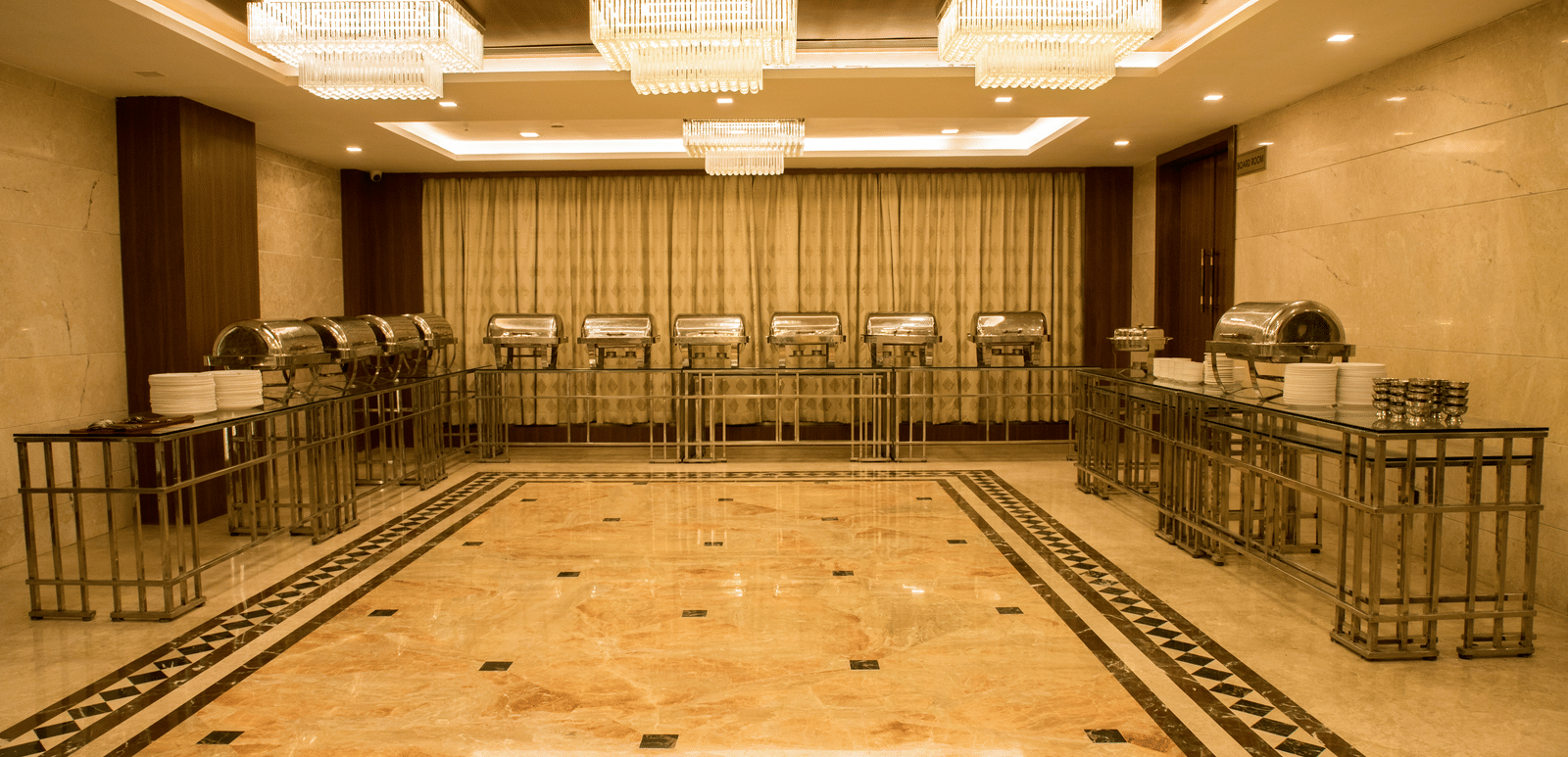 A brightly lit banquet hall with several chafing dishes lined at the far end of the hall at Benzz Park, Vellore.