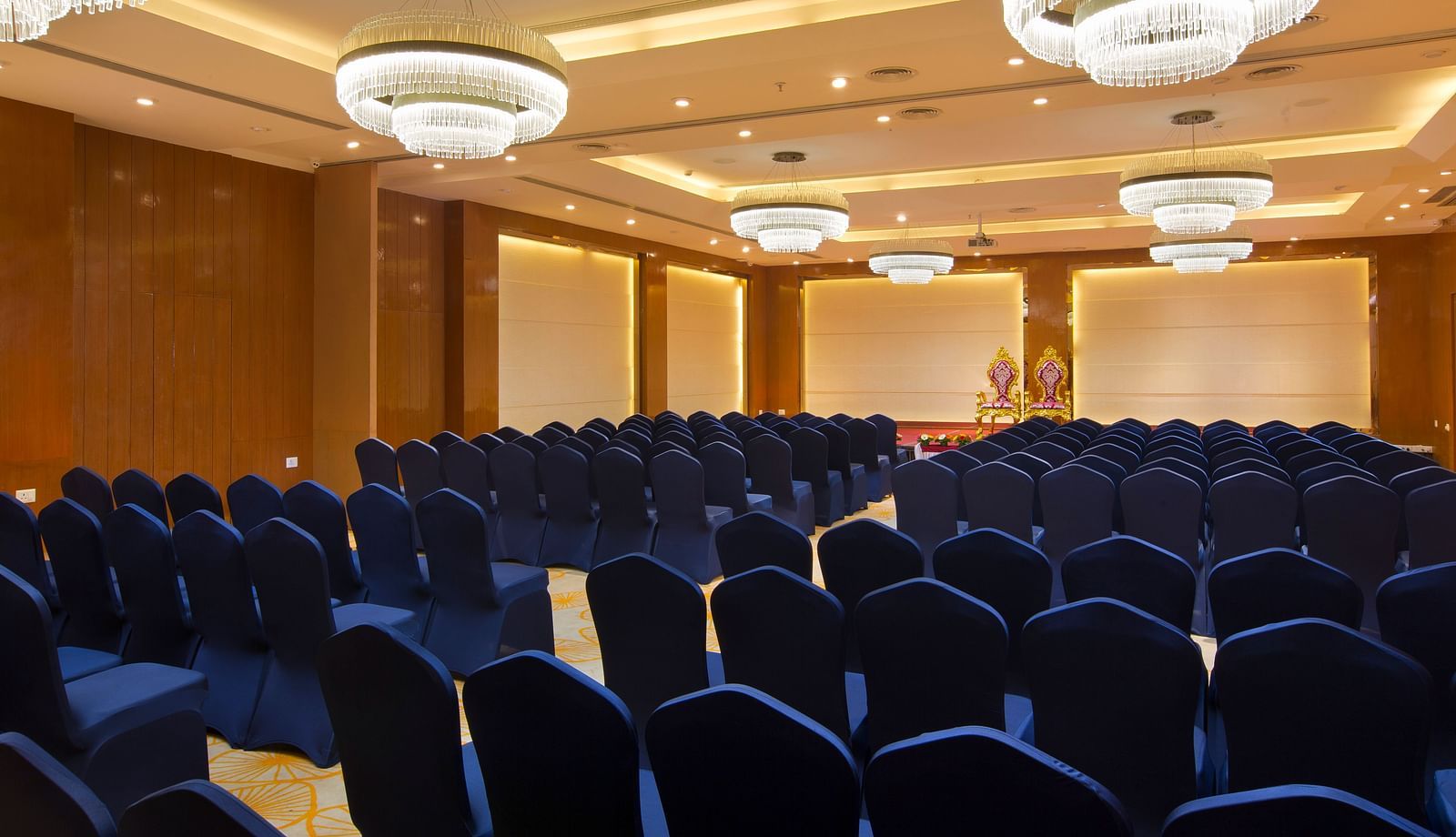 Another view of a spacious conference hall with rows of blue chairs facing a stage with golden figurines, illuminated by large chandeliers - Hotel Southend by TGI - Bommasandra, Bangalore