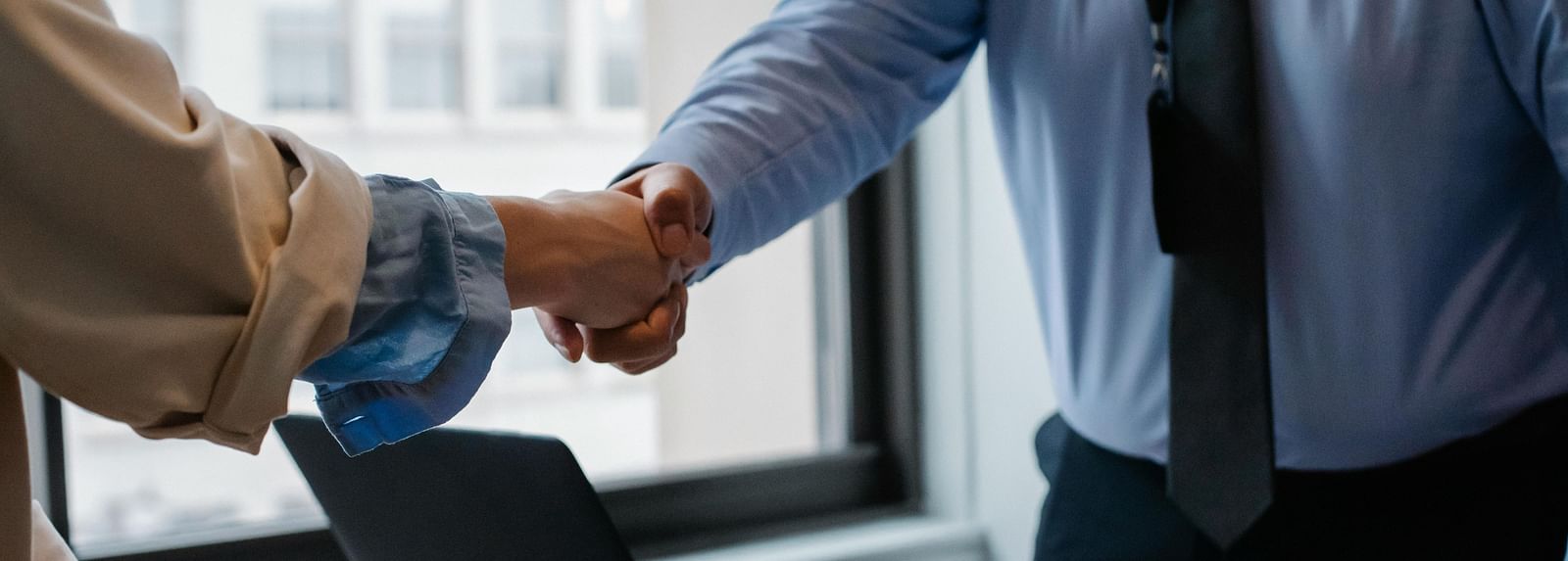 A person dressed in business formals going for a handshake with a laptop and a notepad kept on the table.