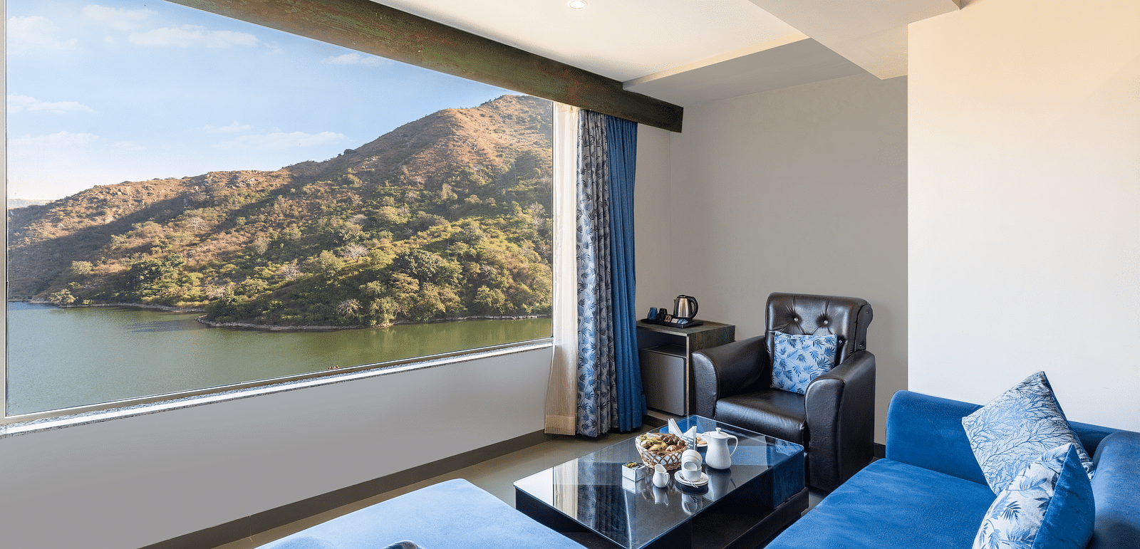 Living area with a blue couch and a large window with a view of mountain and water - Via Lakhela Resort & Spa