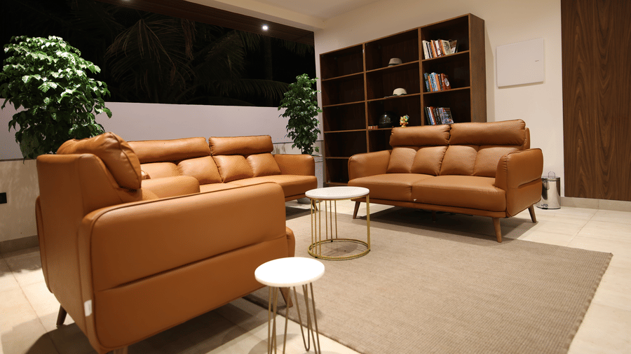 A living area at SUKAVI, featuring 2 leather sofas, small round side tables, a rug, a tall bookshelf, and potted plants.